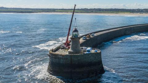 A red crane placing the new dome on relatively short, brick lighthouse. It sits on the circular end of a brick pier which arcs back to the shoreline. The lighthouse is surrounded by white vans and the coast is in the distance.