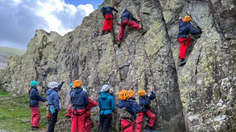 Children wearing protective clothing and helmets climb a steep cliff face