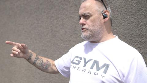 A man with grey hair and beard us pointing to the right, he is wearing a white t shirt. The background is a gravel wall. 