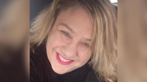 A selfie of Sarah Forrester. She is smiling at the camera and has pink lipstick on. She has shoulder-length, blonde hair and is wearing a black top.