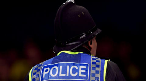 A close-up of the back of a police officer, who is wearing a blue and yellow trimmed vest with the word Police across it. They have a helmet on, and an earpiece in
