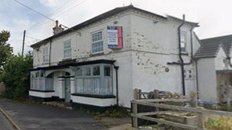 A white building with some marks on the wall with a sign on it. There are large front windows and an arch over the door.