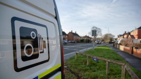 A close up image of a white police camera van. The van has a black image of a speed camera and is facing a nearby road with a car travelling towards it.