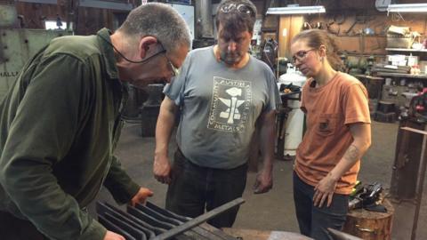 Alan Evans shows two other black smiths some of his work in his Stroud workshop. He wears a green fleece and glasses and the male and female observers wear grey and orange t shirts. It is unclear what the work is.