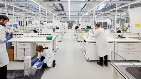 A white laboratory-type building with white work stations where people in white lab coats are working. The work stations have drawers underneath them. A glass roof is held up by white poles.