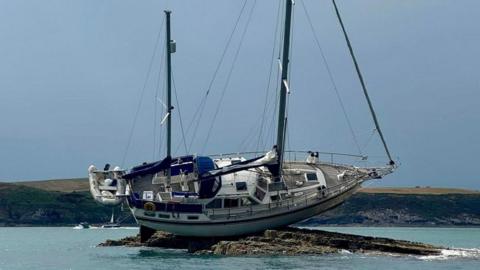 A white yacht on its side on rocks surrounded by sea, with the coastline visible in the background. The yacht was two masts and rigging, with a white dingy attached to it. 