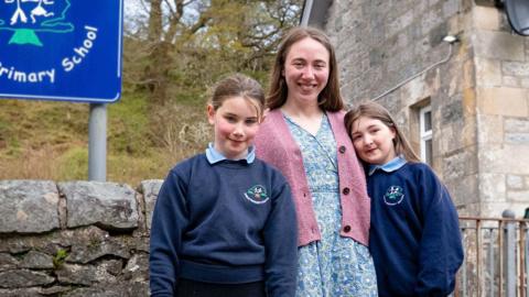 Woman in a pink cardigan with two girls in sweaters on either side