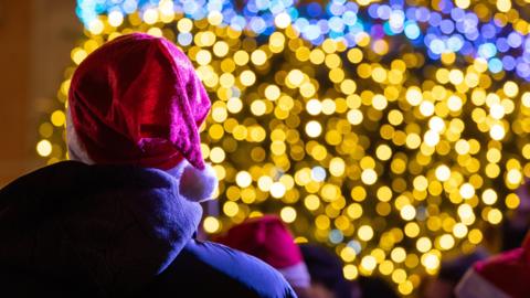 Someone wearing an anorak and a red Santa had with white bobble is looking at a Christmas tree with their back to the camera. The tree is covered with yellow and blue lights but they are out of focus and so create a glow. A shoulder of someone else shows to their right and there is another person wearing a Santa hat in front of them.