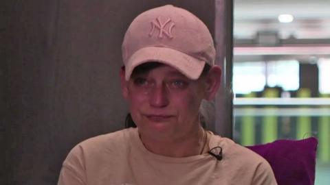 Collette, wearing a pink t-shirt and pink baseball cap, looks into the camera with tears in her eyes 