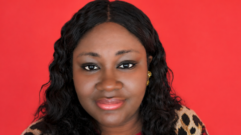 A woman with long dark hair is prominent in the photo and looking at the camera. The plain background behind her is entirely red.