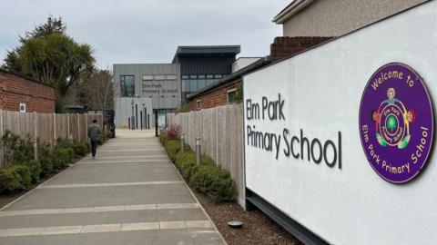 At the entrance to the school, a white wall with silver lettering spells out 'Elm Park Primary School' next to a round purple and yellow logo featuring children holding hands in a circle. A row of wood fencing leads towards a modern grey panelled building with another silver sign for the school