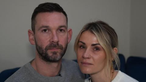Ryan and Kimberly Horsfall sit on a sofa and look at the camera. Ryan has short dark hair and a dark beard. He wears a grey jumper. Kimberly has blonde hair that has been tied up behind her head, with some bangs that frame her face. She is wearing a white jumper. 