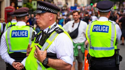 Three police officers stand in high visibility vests in a crowd of people as one man dressed in summer clothes approaches them