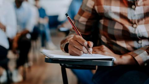 A man sitting in a exam hall with his peers sat around him. He is sitting on a chair with the desk connected. He is in a exam while wearing a plaid button-up shirt.