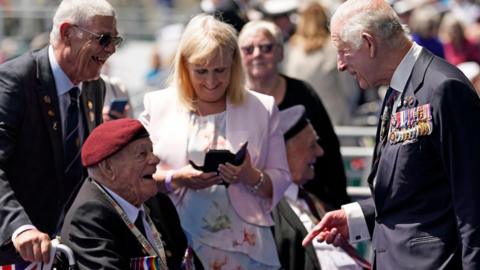 King Charles III talks to WW2 war veteran who is in a wheelchair. It is at n event to commemorate the 80th anniversary of D-Day. The man in the wheelchair has a big smile on his face and is wearing a suit with a red beret.  King Charles is also wearing a suit, both men have their military medals displayed on their jackets. 