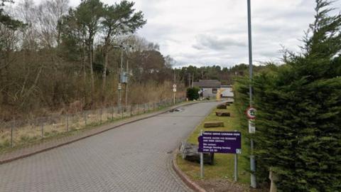 A paved entrance way to a council run caravan park with trees down the sides and a 5mph speed limit sign