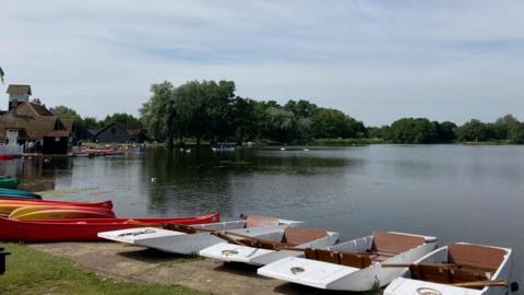 Boats are on the edge of a lake, buildings can be seen on the left edge of the lake with trees on the far side. Some bird can be seen swimming on the water.