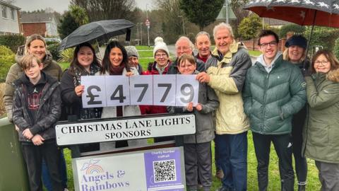 Christmas Lane residents stand in a group and smile at the camera next to a sign that reads: "Christmas Lane Lights Sponsors". Some are holding pieces of paper showing the figure £4,779 - the money raised by the display. Some of the residents hold umbrellas.