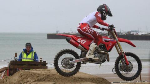 A motocross rider on a red bike in mid air passing over a jump made of sand on Weymouth beach. The rider wears red trousers, w and a white and red top with a black helmet. A safety marshal in a blue coat and a high-viz yellow vest watches from behind a safety barrier.