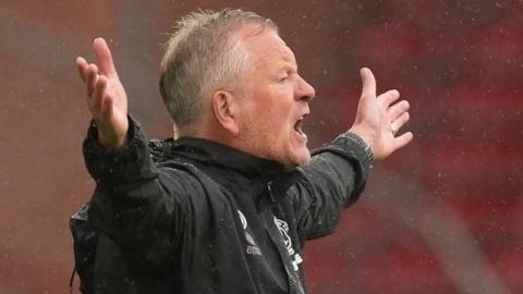 Chris Wilder has his arms outstretched to the side with palms facing forward and opens his mouth in a shout. it is raining and Wilder wears a black rain jacket