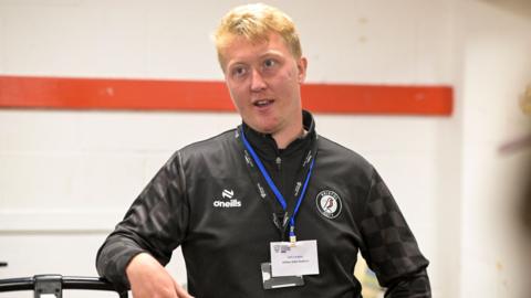 Jack Langley, wearing a black top and a lanyard with his name on it. He is a middle-aged man with ginger hair, and is pictured in a dressing room mid-conversation