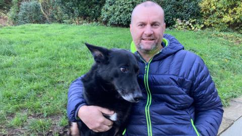 Steve Cockerham, who has shaved hair and grey stubble, poses for a photo with his dog Molly, a black German Shepherd.