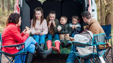 Children sitting together in the boot of a van in the woods eating packed lunch and two adults sitting on camping chairs outside.