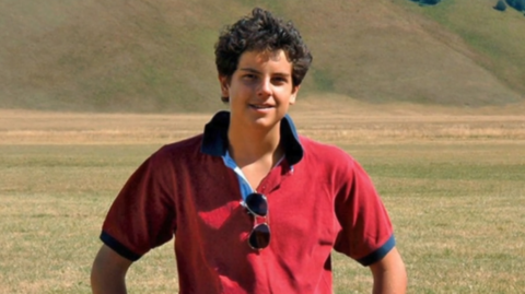 A boy with dark curly hair in a red polo shit stands, smiling at the camera, with his hands on his hips, in front of a field and hills