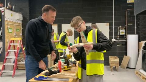 A man in a black top and jeans showing a young man with glasses and wearing a yellow hi-vis jacket some woodworking skills