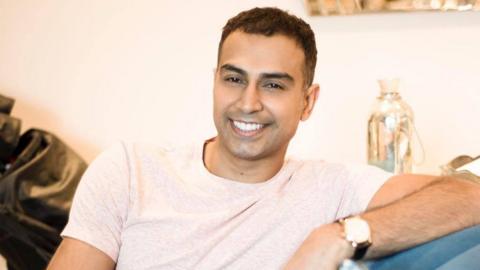 A man who appears to be in his 30s - he is sitting on a sofa at home, wearing a light off-white t-shirt, and smiling, clearly relaxed in the setting - he is leaning on the arm of the sofa with his left arm, and his watch is visible 