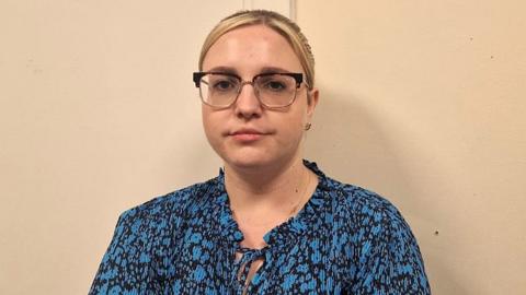 Sophie has blonde hair and wears glasses. She is wearing a blue blouse and looking at the camera. 