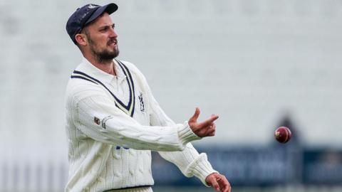 New Warwickshire captain Ed Barnard is tossing a ball underarm to a colleague out of shot, wearing whites with a woollen Warwickshire club jumper and blue club cap.