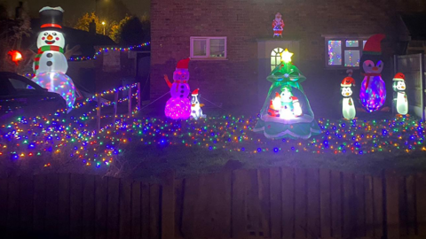 Christmas lights and illuminated inflatables including snowmen and penguins in the garden of a house. Lights are strewn across the lawn, with a fence in the foreground