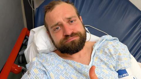 A man with short brown hair and a beard has a sad face as he lies in a hospital bed in a gown. 