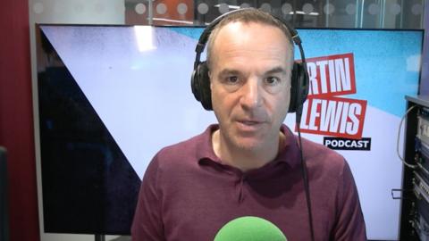 Martin Lewis, wearing a burgundy top, in front of a green microphone, wearing headphones and sitting in from of a large TV