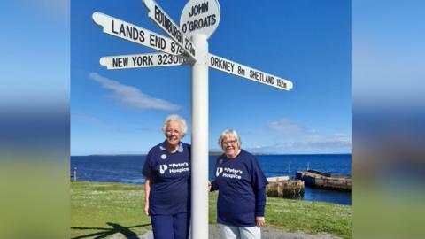 Valerie and Sue are standing next to a white direction sign post in John O'Groats. The post is pointing in multiple directions to New York, Edinburgh and Lands End. 