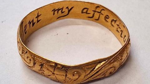 Close up of the gold ring, a band with engraving round the outside and the wording inside that says 'present my affection'