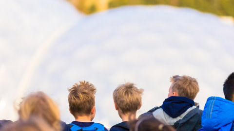 The picture shows a group of young people seen from behind, standing close together outdoors. Several are wearing backpacks and padded jackets in shades of blue, grey and black, suggesting cool weather. The focus is on the backs of their heads and shoulders, with short hair visible on a few individuals. In front of the group is a large, pale, rounded surface of the Eden Project biomes.