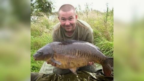 Terry Ricketts wearing a green t-shirt and trousers kneeling down in the grass holding a large fish and smiling at the camera