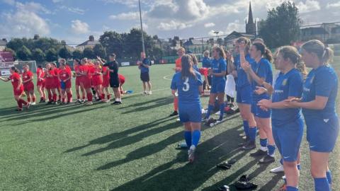 There are two teams on the pitch, the Jersey team is wearing a blue strip and are clapping and the other team is wearing a red strip