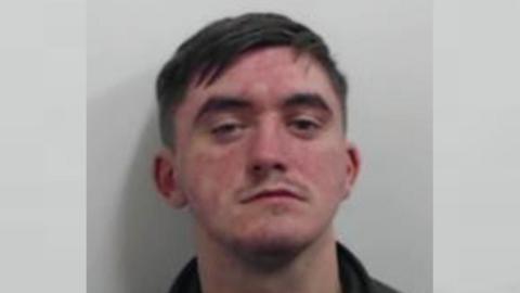 A mugshot of a man with brown hair and a faint moustache. He is wearing a black top.