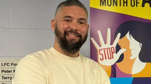 Tony Bellew, 43, who has dark hair speckled with grey and a large black beard, smiles at the camera in front of a sign with slogans including 'Schools Month of Action' and a cartoon image of a woman holding up her hand with the word 'Stop' written on her palm in red lettering. 