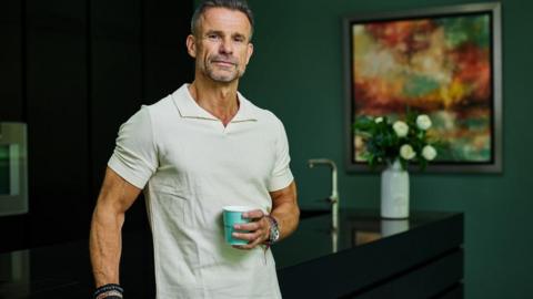 A man with greying hair in a white polo shirt. He is wearing a watch and some bracelets. He is holding a aqua-coloured cup and leaning on a kitchen counter.