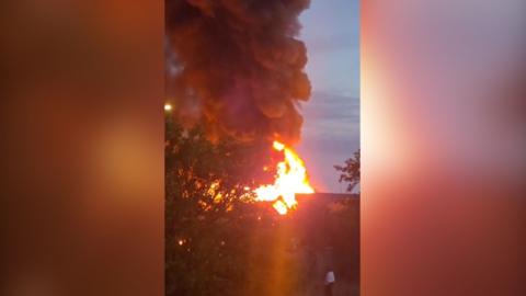 Thick plumes of smoke and flames rising into dawn sky from warehouse fire