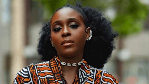 Portrait of a woman standing outdoors in a black, white, and orange patterned garment with a shell necklace and decorative ear cuff. Trees and buildings are softly blurred in the background.