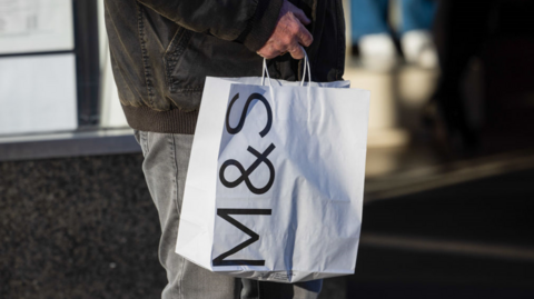 Man in dark winter jacket and grey jeans carries white paper bag with M&S logo on the side