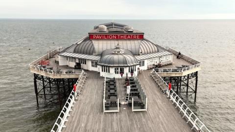 A long wooden pier extends over calm sea water toward a building labelled “Pavilion Theatre.” White railings line both sides of the pier - with benches on the decking.