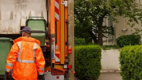 A bin crew member wearing an orange jacket and a black cap- standing in front of an orange lorry emptying green bins