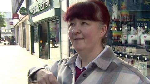 A woman with dark hair, dyed slightly red, looks to the left of the camera. She stands in front of the glass windows of a shop with a sign for "Holland and Barrett" over a shop next to that. She has her hair pulled back behind her head and wears a light checked shirt over a mauve top