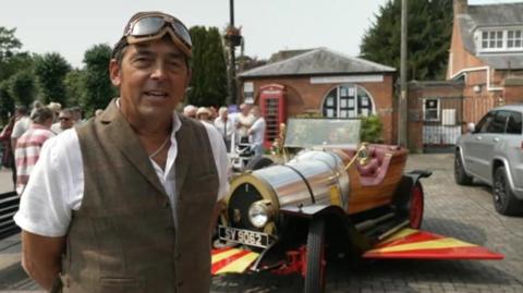 A man in a waistcoat and driving goggles standing next to a replica of Chitty Chitty Bang Bang